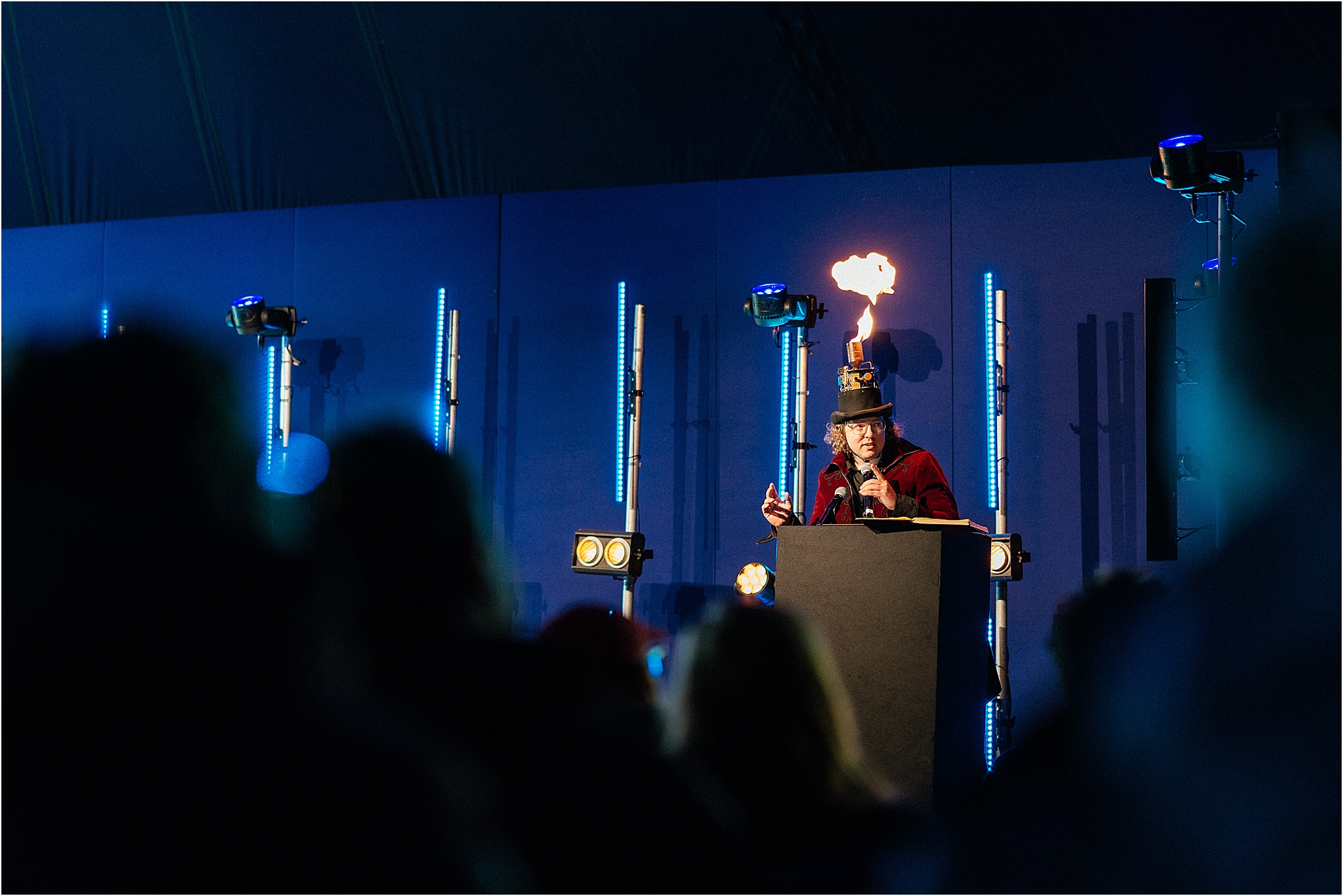 A photograph of a speaker giving a talk with a flaming hat at a festival. 
